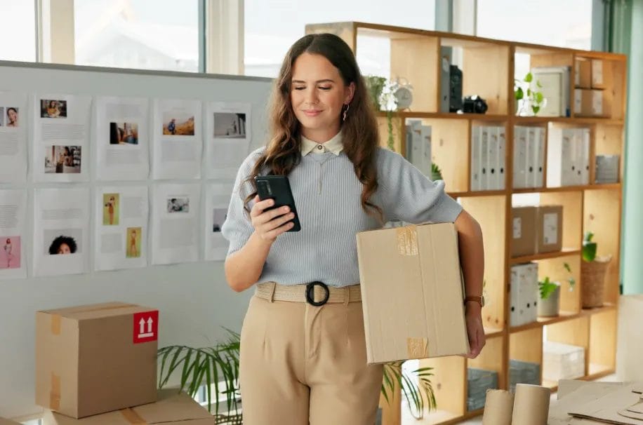A small business owner wearing a light blue sweater and khaki pants smiles while checking her smartphone and holding a boxed package for shipment in her studio with a whiteboard and wooden shelving in the background.