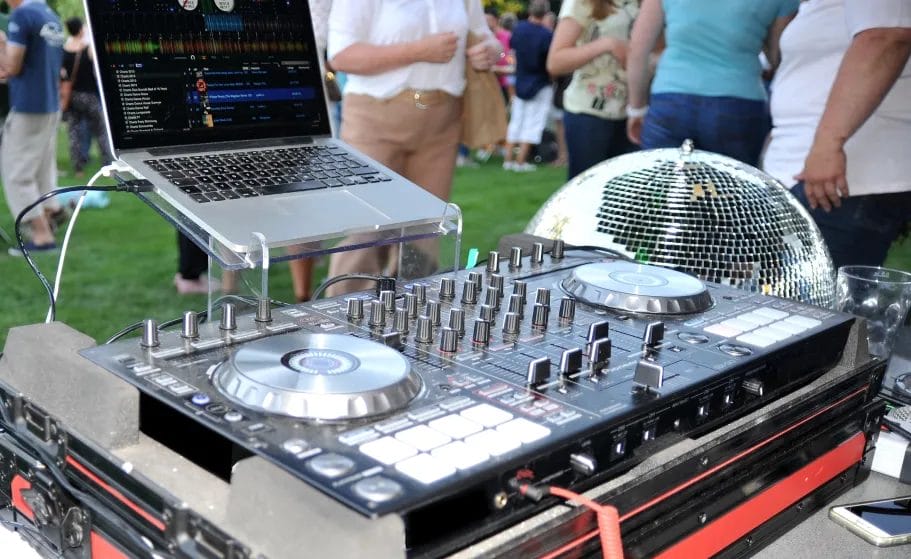 A close-up image of a professional DJ's equipment setup, including a laptop, mixing console, and a disco ball at an outdoor event with green grass and attendees in the background.