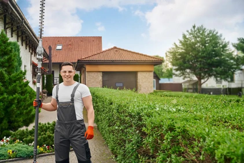 A landscaping professional wearing a white shirt, gray overalls, and red gloves smiles while holding an electric hedge trimmer outside of a client's home beside a hedgerow.