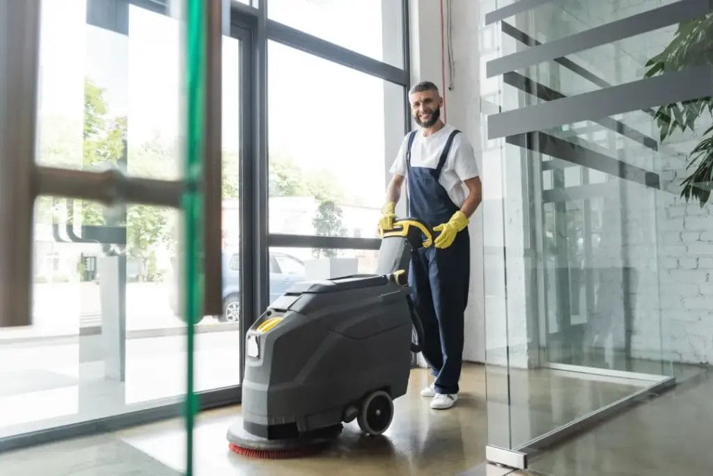 A cleaning business owner wearing a white shirt, navy overalls, and yellow gloves smiles while cleaning a commercial building floor with an automatic floor scrubber machine.