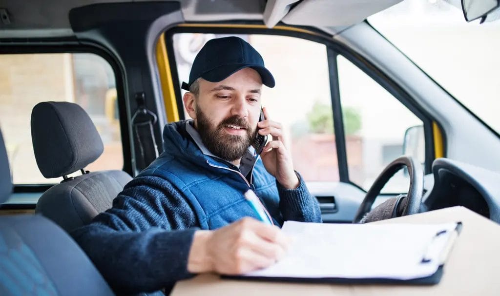 A business owner sits in the driver's seat of a parked van where he speaks on the phone and reviews paperwork for a delivery.