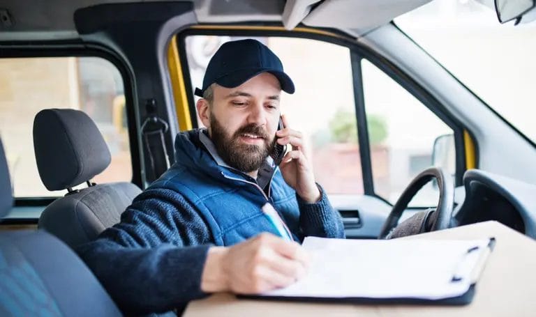 A business owner sits in the driver's seat of a parked van where he speaks on the phone and reviews paperwork for a delivery.