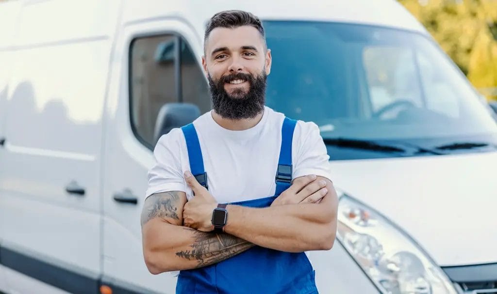 A businessman in a white tee and blue work overalls smiles with his arms crossed as he stands in front of a work van.