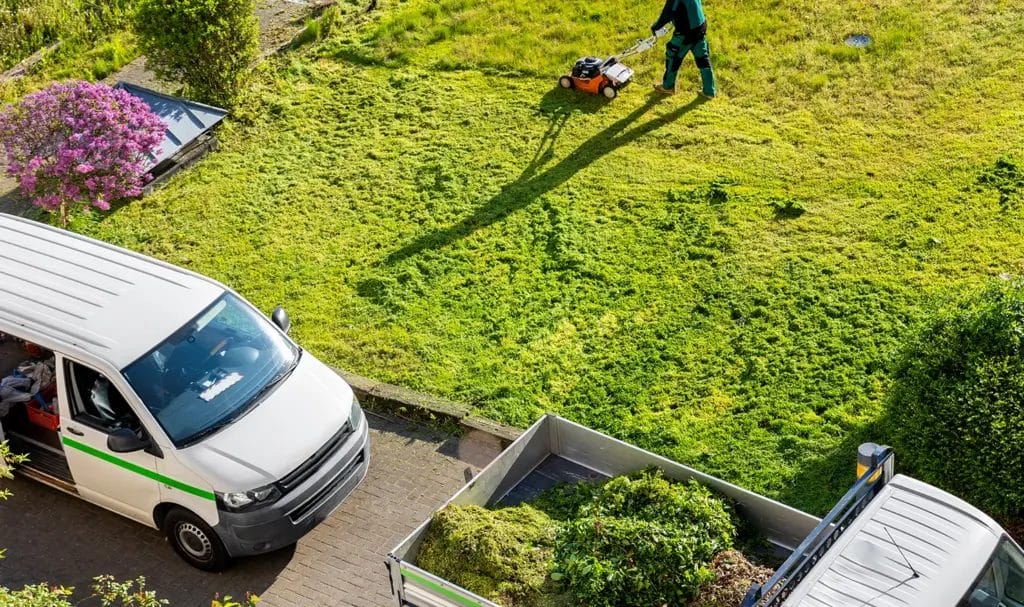 An overhead view of a landscaper mowing a lawn, while a work van and truck loaded with supplies are parked on the driveway near him.