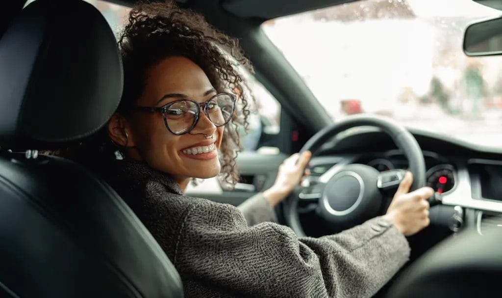 A young woman turns back to smile from the driver's seat of a car she's driving around for work.