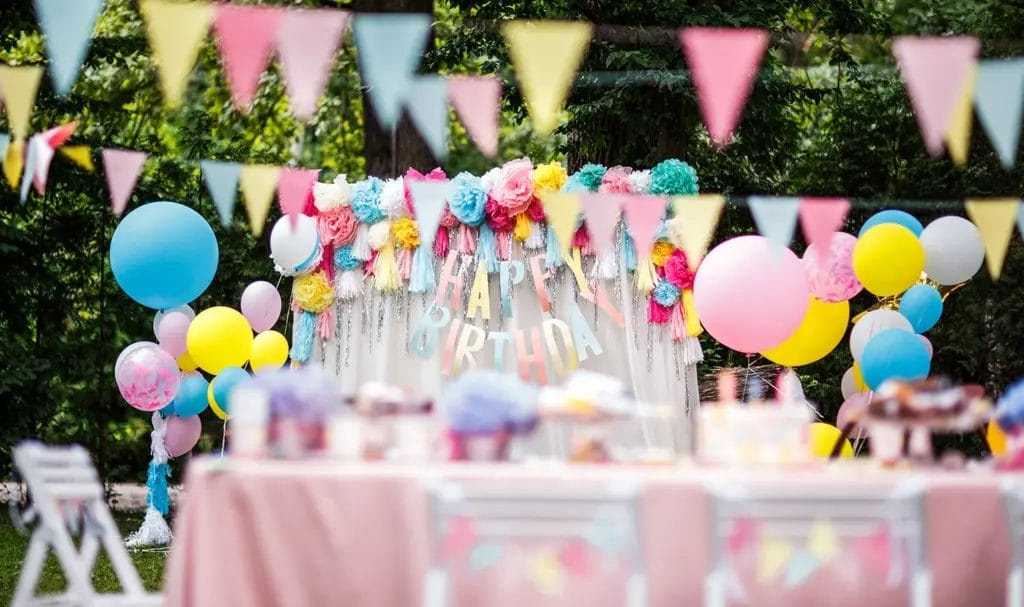 A backyard birthday party setup, complete with colorful banners, balloons, tablecloths, and a photo backdrop with a banner that says "Happy Birthday."