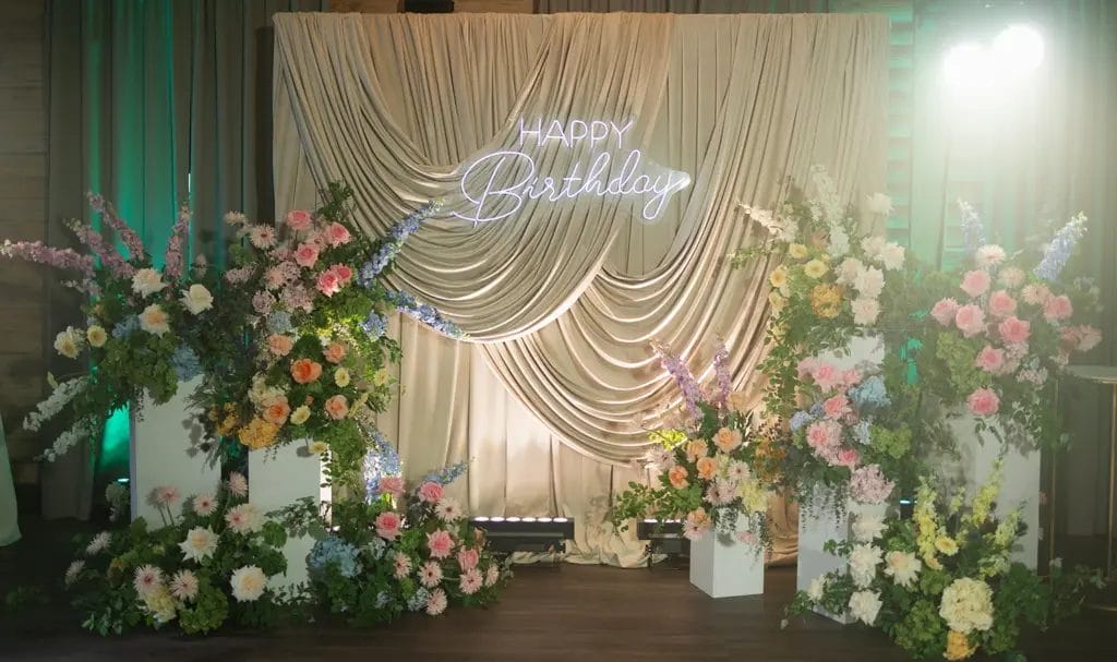 An elegant backdrop of draped fabric and various, colorful florals on pedestals, with a neon sign that says "Happy Birthday."