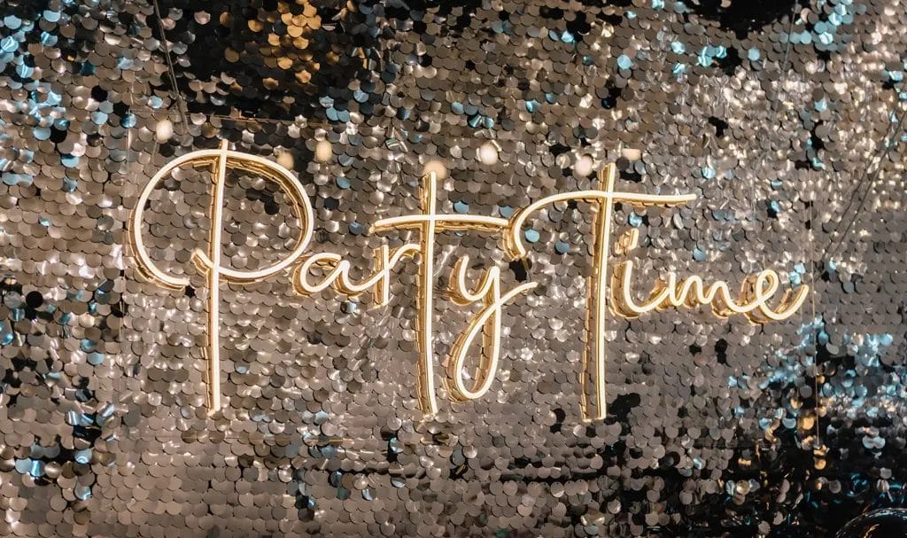 A neon sign that reads "Party Time" sits against a silver sequined backdrop.