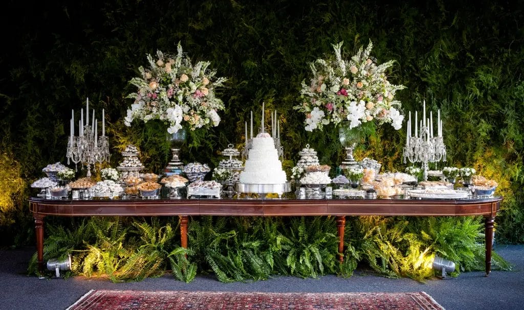 An elegant wedding dessert table with florals sits against a green fern backdrop wall.