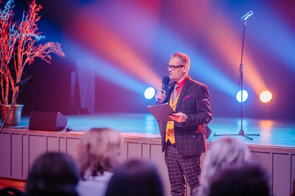 a male event host speaking into a microphone in front of an audience seated before a stage