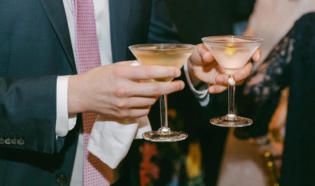 Close-up of a man in a suit and pink tie carrying two martinis through a crowded wedding party.
