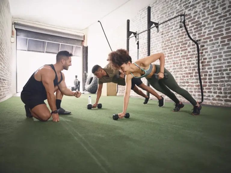 Instructor, dumbbell and plank with people in gym for fitness, health and challenge. Weightlifting, coaching and exercise club with group in sports center for workout, personal trainer and class