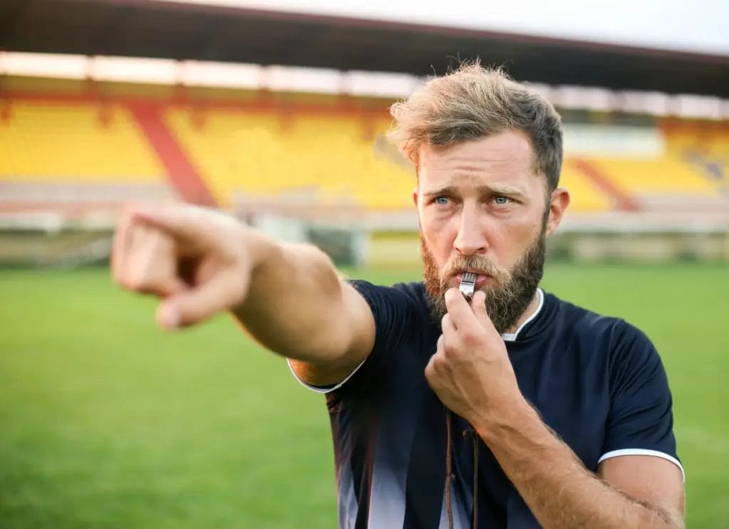 A football referee with a beard points and blows his whistle