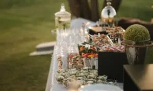 A side-shot of a table with a white tablecloth and catered foods and beverages arranged for an outdoor wedding.