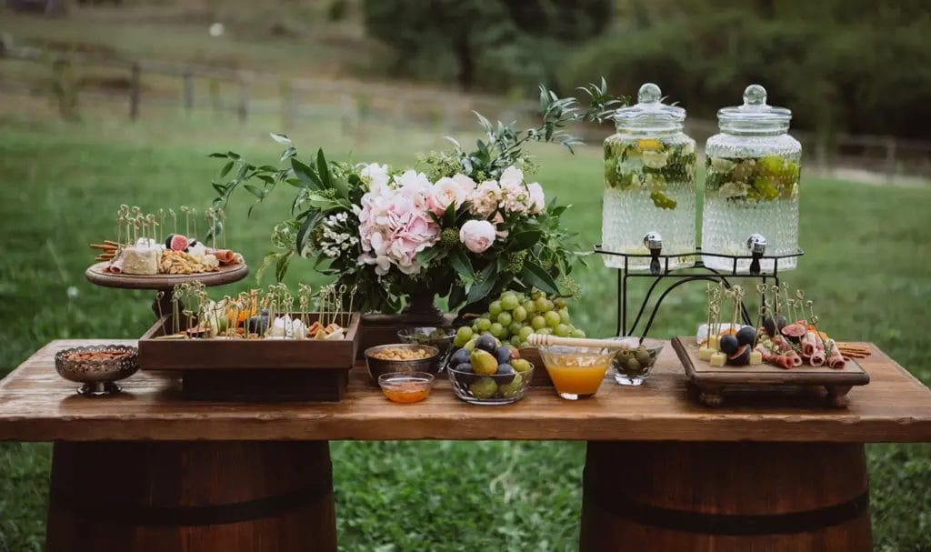 A wood tabletop supported by two wine barrels has an array of catered food and beverages beautifully displayed for an outdoor event hosted at a winery.