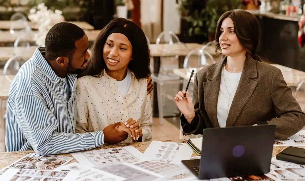 A wedding event planner speaks with a couple about plans for their upcoming wedding, with a laptop and inspiration photos laid out on the table in front of them.