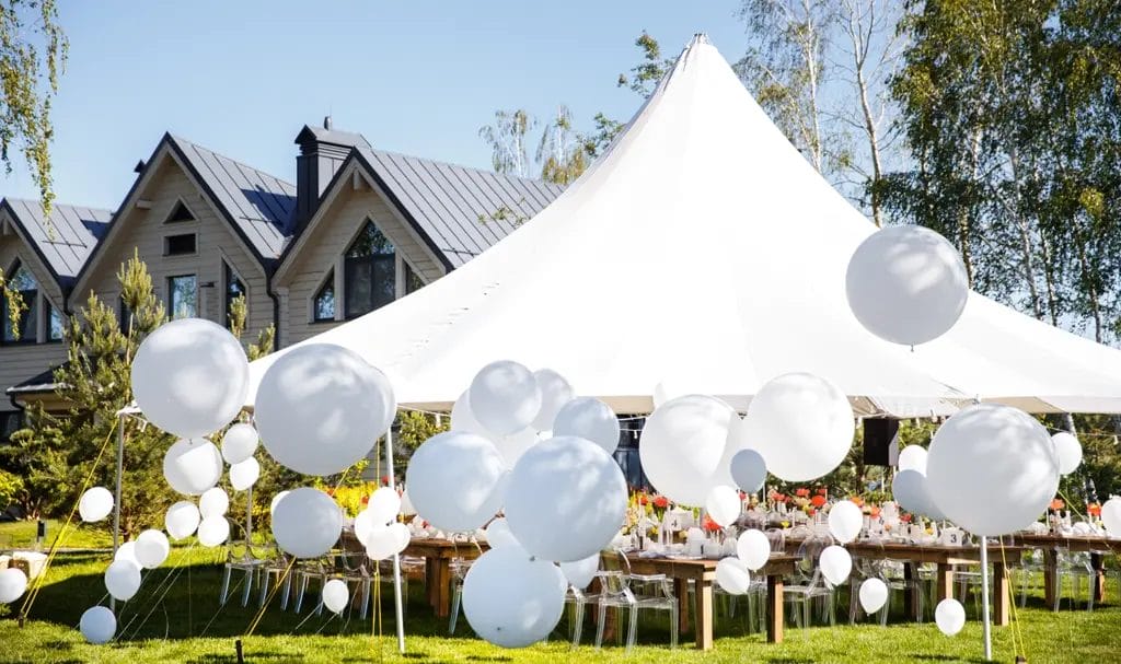 A white event tent covers rows of decorated tables and white balloons, set up for a celebration on the lawn of a private residence.