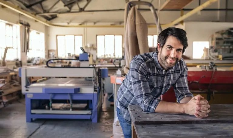 Man in workshop smiles as he leans forward on to a workbench.