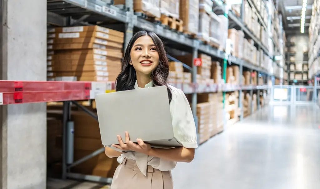 A businesswoman smiles while holding her open laptop and walking down the aisle of a warehouse where she checks produce inventory.