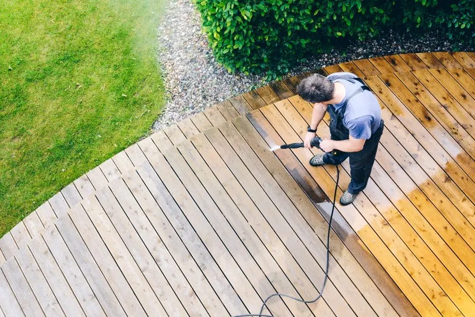 A power washing professional wearing a light blue shirt and navy overalls power washes a wood-slat outdoor platform in a garden with gravel, grass, and shrubbery.