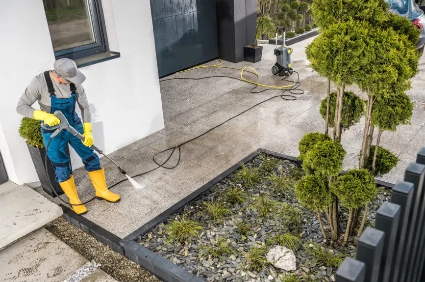 A pressure washing professional wearing blue overalls and yellow boots pressure washes a walkway outside of a property decorated with green trees and shrubbery.