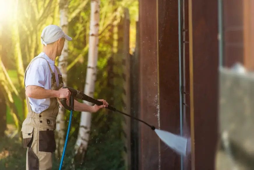A pressure washing professional wearing a white hat, white shirt, and khaki overalls pressure washes the side of a brown building.