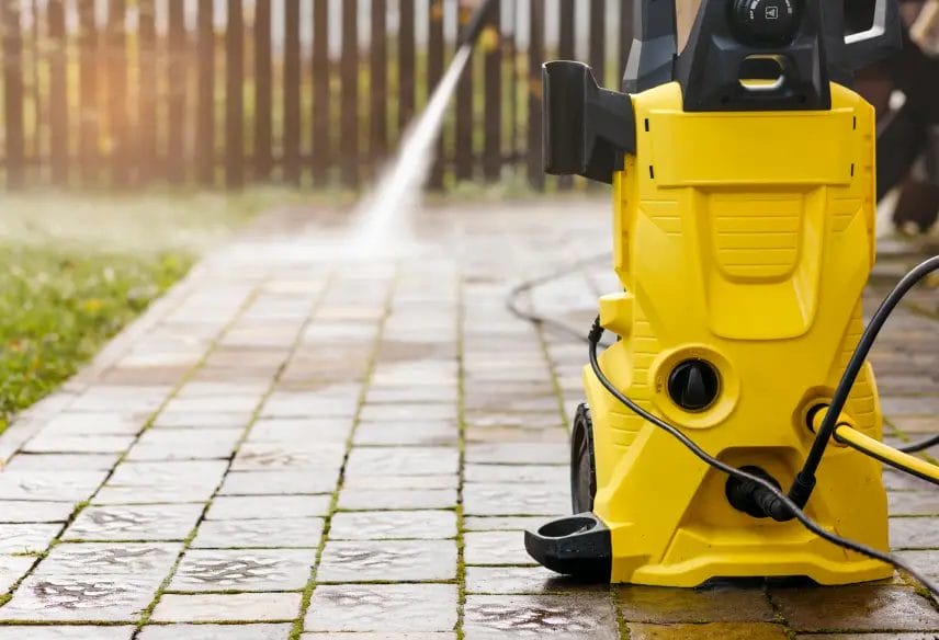 A close-up image of a yellow pressure washer set on an outdoor tiled floor, with the spray from the machine cleaning the floor in the background.