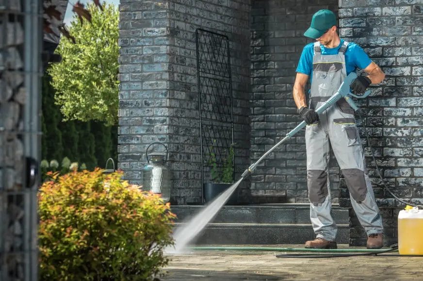 A pressure washing professional wearing a green hat, blue shirt, and gray overalls pressure washes a walkway outside of a blue building with shrubs and trees decorating the property.