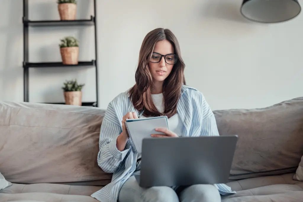 Woman wearing glasses, sitting on a couch with a notebook and a laptop.