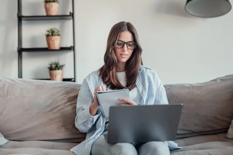 Woman wearing glasses, sitting on a couch with a notebook and a laptop.